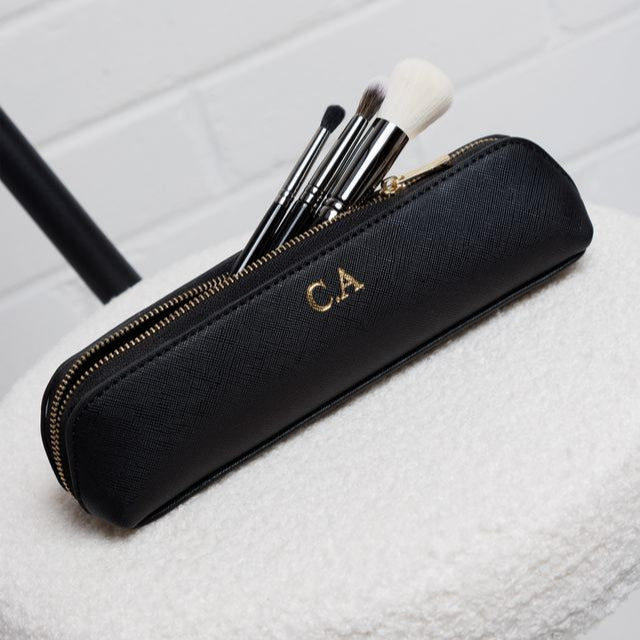 Black makeup brush set with gold initials on a white stool against a white brick wall.